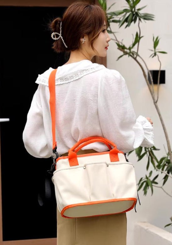 Une femme aux cheveux bruns coiffés en chignon bas porte un chemisier blanc bordé de dentelle et une jupe kaki. Elle porte le Sac a langer | Bébé Longchamps, un sac à bandoulière orange et blanc, à côté de plantes vertes d'intérieur.