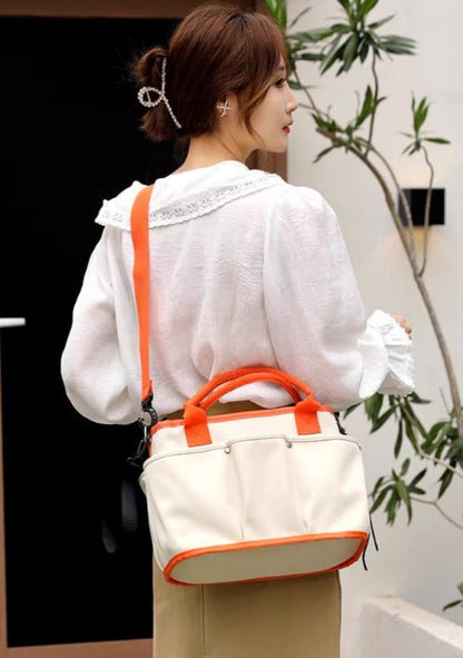 Une femme aux cheveux bruns coiffés en chignon bas porte un chemisier blanc bordé de dentelle et une jupe kaki. Elle porte le Sac a langer | Bébé Longchamps, un sac à bandoulière orange et blanc, à côté de plantes vertes d'intérieur.