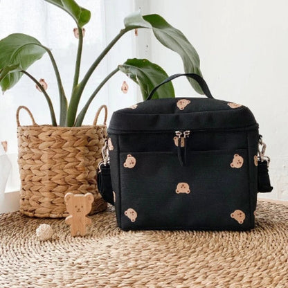 A Sac a Langer Bebe - TeddyBear Organizer avec un adorable visage d'ours repose sur un tapis tissé à côté d'une plante en pot et d'un ours en bois. Le sac est muni d'une poignée, d'une poche frontale et d'une bandoulière pour plus de commodité.