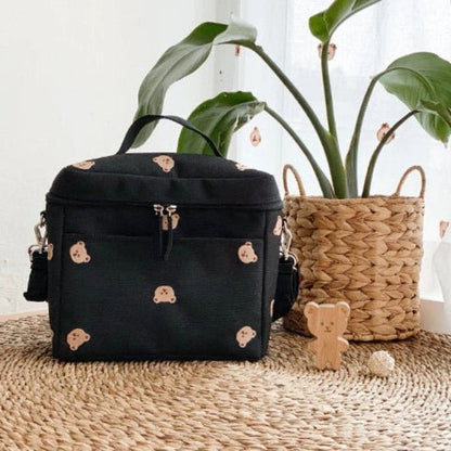 Le Sac a Langer Bebe - TeddyBear Organizer, avec ses petits visages d'ours, est placé sur un tapis tissé à côté d'une plante en pot dans un panier et d'une figurine d'ours en bois, avec des rideaux blancs transparents en arrière-plan.
