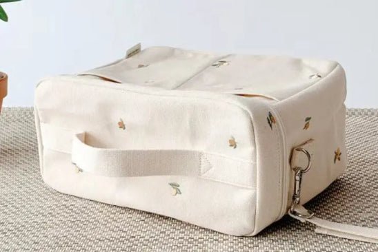 Le Sac à Langer Bébé, un sac rectangulaire en tissu crème avec anse, repose sur une surface texturée beige. Il est doté de petits motifs brodés et d'un clip en métal, offrant une protection optimale. Une plante partiellement visible renforce sa fonctionnalité et son style.