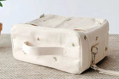 Le Sac à Langer Bébé, un sac rectangulaire en tissu crème avec anse, repose sur une surface texturée beige. Il est doté de petits motifs brodés et d'un clip en métal, offrant une protection optimale. Une plante partiellement visible renforce sa fonctionnalité et son style.