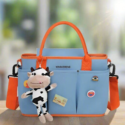 Le Sac à langer | Bébé Longchamps est un sac à langer bleu clair avec des poignées et des bretelles orange, orné d'un porte-clés vache en peluche, de pin's et d'une carte postale. Il est posé sur une surface en bois sur un fond flou.