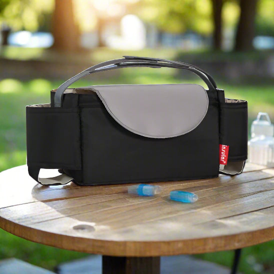 A Sac à langer bébé - Organisateur poussette, noir et gris et isolé, est posé sur une table d'extérieur en bois avec deux pains de glace bleus et un biberon à proximité. La lumière du soleil filtre à travers les arbres verts, parfaits pour des sorties sereines.