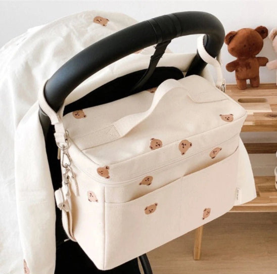 Un Sac à Langer Bébé couleur crème, orné de petits visages d'ours, est suspendu à la poignée d'une poussette, alliant praticité et style. Derrière, un ours en peluche repose sur une étagère en bois.