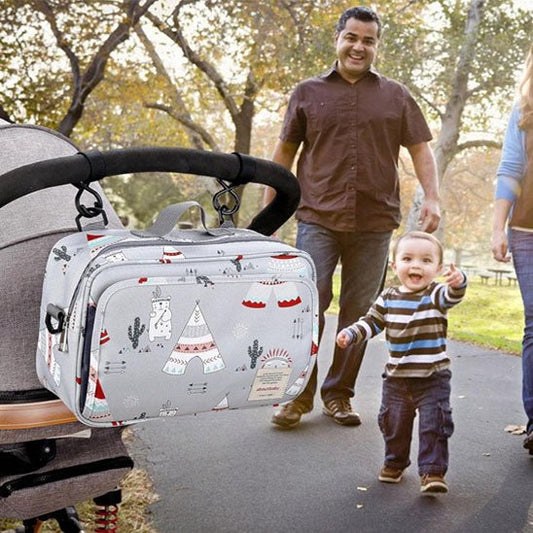Un sac à langer bébé | Une poussette grise avec un imprimé tipi et ours est suspendue à la poignée d'une poussette, tandis qu'un couple souriant et un tout-petit se promènent sur un chemin de parc ensoleillé et bordé d'arbres - des accessoires de bébé parfaits pour les sorties.