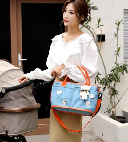 Une femme en chemisier blanc se tient à côté d'une poussette beige, tenant le Sac à langer | Bébé bleu et orange, orné de peluches et de pins, indispensable pour les parents en déplacement. Des plantes et une porte sont visibles en arrière-plan.