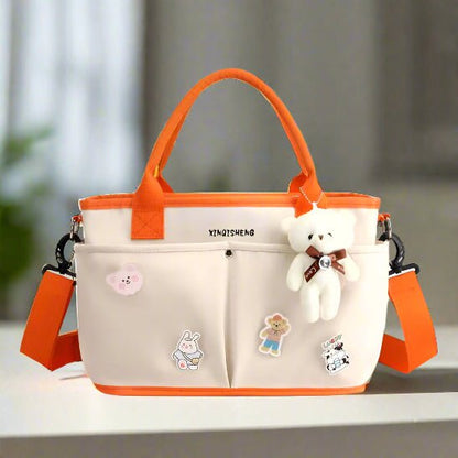 Le Sac à langer | Bébé Longchamps, un sac à langer beige et orange avec des poignées orange, une sangle, des épingles de bande dessinée et un porte-clés en forme d'ourson blanc, repose sur une surface claire sur un fond d'intérieur légèrement flou.