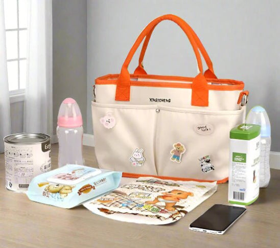 Le Sac à langer | Bébé beige avec poignées orange est posé sur une table, entouré d'articles essentiels pour bébé comme des boîtes de lait maternisé, des biberons, des lingettes, un bavoir, des mouchoirs, une boîte de jus et un smartphone, parfait pour les parents en déplacement.