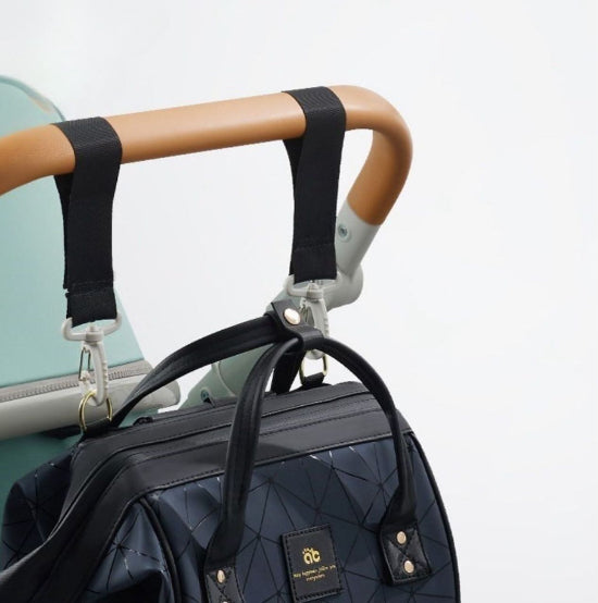 Un sac à langer noir matelassé est suspendu au guidon marron clair d'une poussette avec des sangles élégantes, idéal pour les parents modernes. La poussette bleu clair chic complète le sac, qui comporte une petite étiquette et des fermoirs métalliques sur un fond blanc impeccable.