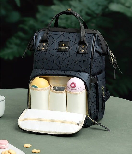 Le Sac à Langer dos, un sac à langer noir au motif géométrique, est ouvert pour dévoiler trois poches isothermes contenant des biberons ronds. Idéal pour les parents modernes en sortie familiale, il repose sur une table verte avec une tasse en partie visible et des biscuits éparpillés sur un fond de verdure floue.