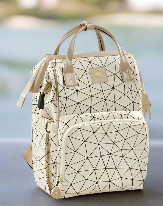Le Sac a Langer dos | LifeEscape™ est un sac crème avec des lignes géométriques noires, des fermetures éclair dorées, deux poignées et une poche frontale zippée - idéal pour les parents modernes - présenté sur une surface grise avec un flou extérieur en arrière-plan.