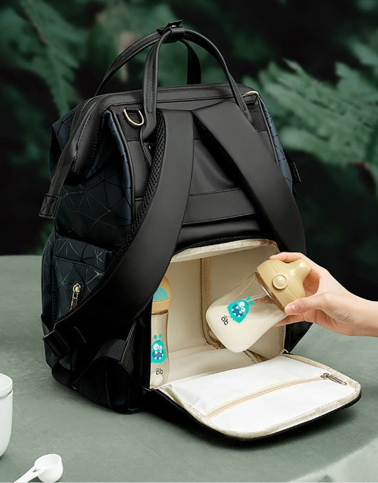 Le Sac à Langer dos, un sac à langer noir au design matelassé, est posé ouvert sur la table. Il est doté d'un compartiment avant isolé pour les biberons. Une main ajuste un biberon au bouchon beige sur un feuillage vert légèrement flou. Parfait pour les parents modernes !.
