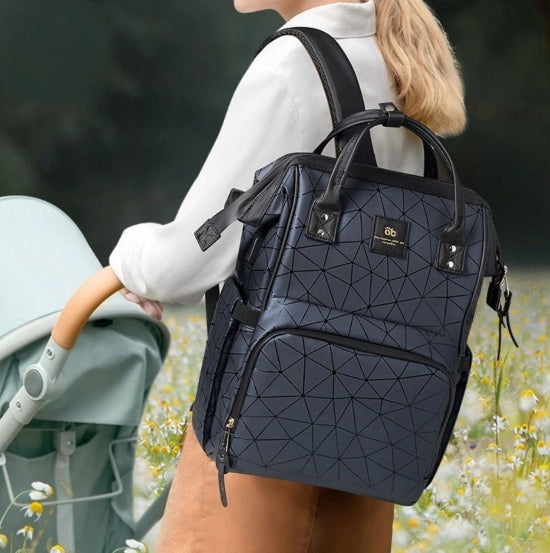 Une personne aux cheveux longs et clairs porte un Sac à Langer dos à motif géométrique tout en poussant une poussette verte claire dans un champ de fleurs. Le sac à dos foncé avec bretelles noires et petit logo est parfait pour les parents modernes en sortie en famille.