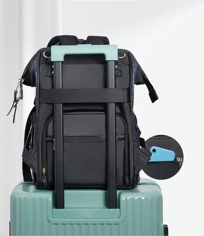 Un Sac à Langer dos, un sac à dos noir à fermetures éclair multiples et à poignée accentuée bleu clair, idéal pour les parents modernes, est fixé à la poignée d'une valise bleu sarcelle. Une pochette circulaire dotée d'une tirette de fermeture éclair bleu clair complète le design. La scène se déroule sur fond de rideau blanc.
