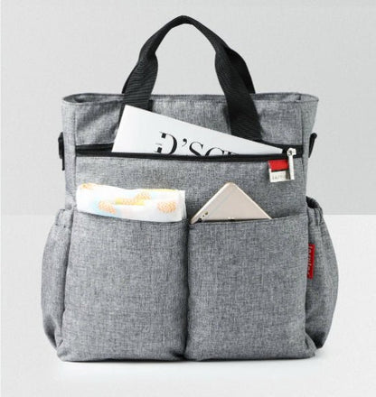 Le Sac à Langer - Fourre-tout, un cabas en tissu gris aux poignées noires, repose sur une surface blanche. Ses poches avant permettent de ranger un smartphone et un chiffon, tandis que le compartiment principal accueille un magazine : idéal pour les parents modernes.