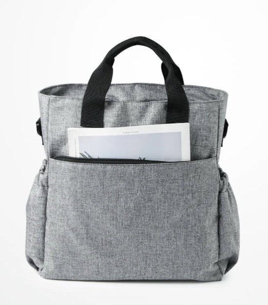 Le Sac à Langer - Fourre-tout, présenté en tissu gris avec des poignées noires sur fond blanc, est doté de poches latérales pratiques et d'une poche avant partiellement dézippée révélant un journal, idéal pour les parents modernes.