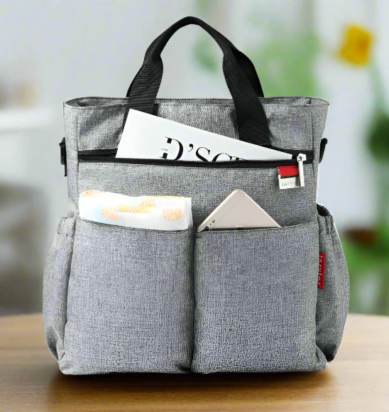 Le Sac à Langer - Fourre-tout, un cabas en tissu gris aux poignées noires, repose sur une surface blanche. Ses poches avant permettent de ranger un smartphone et un chiffon, tandis que le compartiment principal accueille un magazine : idéal pour les parents modernes.