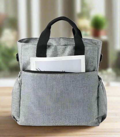 Le Sac à Langer - Fourre-tout, présenté en tissu gris avec des poignées noires sur fond blanc, est doté de poches latérales pratiques et d'une poche avant partiellement dézippée révélant un journal, idéal pour les parents modernes.