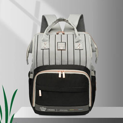 Le Sac à langer - LeQueen Bag pour Bébé est un sac à dos thermique bicolore avec un dessus gris clair à rayures verticales, une poche frontale noire, des fermetures à glissière et des poignées beiges, et un écusson logo. Idéal pour les parents organisés, il est posé sur une étagère blanche.