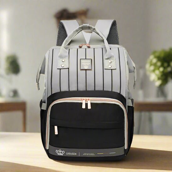 Le Sac à langer - LeQueen Bag pour Bébé, avec ses tons gris et noirs, ses rayures verticales sur le dessus, sa poche zippée sur le devant, ses poignées beiges et ses fermetures éclair dorées, est posé sur une table - idéal pour les parents organisés.