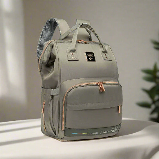 Le Sac à langer - LeQueen Bag pour Bébé, de couleur gris clair avec des fermetures éclair et des poignées de couleur beige, repose sur une surface blanche. Conçu pour les parents organisés, il comporte plusieurs compartiments et une étiquette frontale noire. Une plante apparaît en arrière-plan.