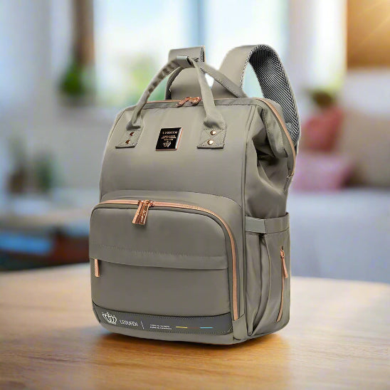 Le Sac à langer - LeQueen Bag pour Bébé, de couleur grise avec des fermetures éclair et des accents de couleur beige, se tient debout sur une table. Il est doté d'une poche frontale, d'un logo noir, de poignées courtes et de bandoulières pour faciliter l'organisation des parents.