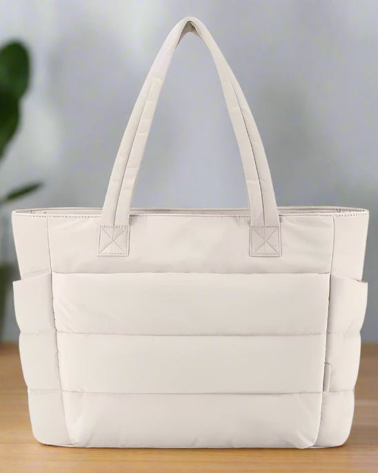 Le Sac a langer | Sac de Maternité Maman, un grand sac à langer beige clair personnalisable avec deux poignées rembourrées, des poches latérales et une forme rectangulaire, repose sur une surface en bois sur un fond flou.
