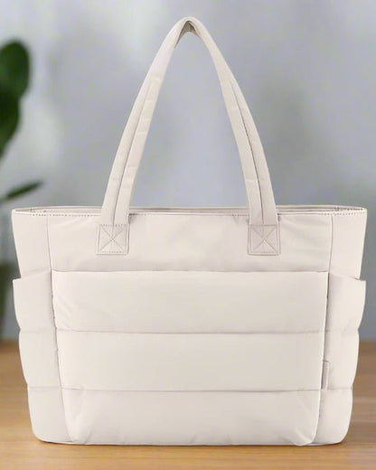 Le Sac a langer | Sac de Maternité Maman, un grand sac à langer beige clair personnalisable avec deux poignées rembourrées, des poches latérales et une forme rectangulaire, repose sur une surface en bois sur un fond flou.