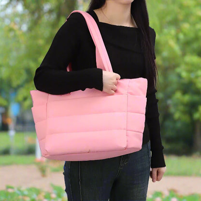 Une personne vêtue d'un haut noir à manches longues et d'un jean se tient dehors, le Sac à langer | Maman, un grand sac à langer rose clair, en bandoulière. Des arbres verts et un parc sont visibles en arrière-plan.
