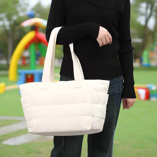 Une personne portant un haut à manches longues noir et un jean tient un « Sac à langer | Maman » blanc matelassé par ses poignées alors qu'elle se tient debout à l'extérieur sur l'herbe avec des équipements de jeux en arrière-plan.