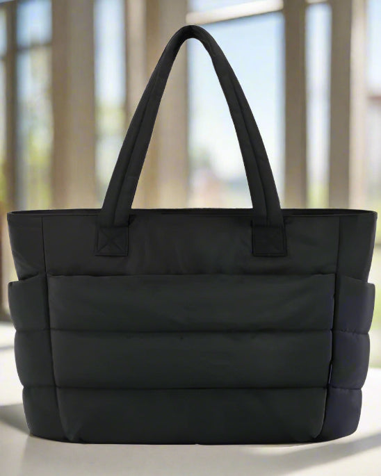 Le Sac à langer | Maman est un grand sac fourre-tout multifonction noir avec panneaux horizontaux matelassés et rembourrés et deux poignées, présenté sur un fond blanc uni.