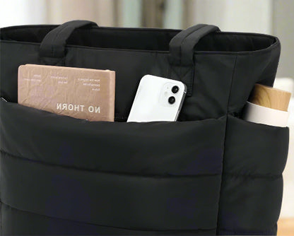 Un sac fourre-tout multifonction noir Sac à langer | Maman avec une poche extérieure contenant un livre, un smartphone blanc et une bouteille blanche avec un couvercle en bois.