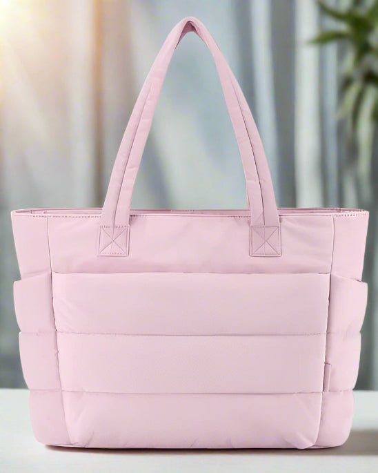 Le Sac à langer | Sac de Maternité Maman, un sac à langer matelassé rose clair avec deux anses, est présenté sur un fond doucement éclairé, avec des rideaux et une plante visibles sur la droite.