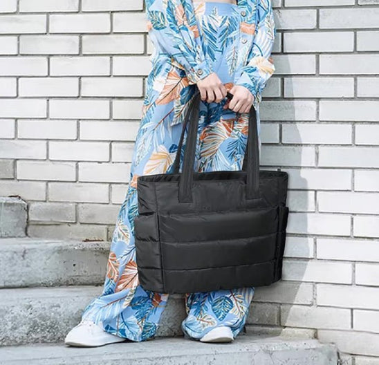 Une personne vêtue d'une tenue bleue à motifs de feuilles colorées se tient sur les marches d'un mur de briques blanches, tenant le Sac à langer | Maman. Son visage est invisible.