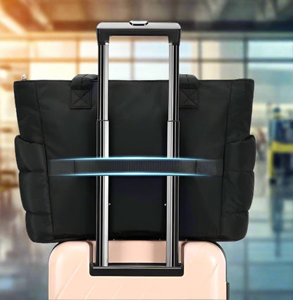 Un Sac à langer | Maman en noir est fixé sur la poignée allongée d'une valise à roulettes de couleur claire, avec sa sangle enroulée sur la poignée de la valise, sur un fond blanc.