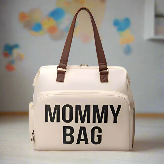 Le Sac à Langer | Mommy Bag, beige avec anses marron et inscription « MOMMY BAG » en gras sur la poche avant, se pose à même le sol. Idéal pour ranger les accessoires de bébé. Un mur coloré et flou est visible en arrière-plan.
