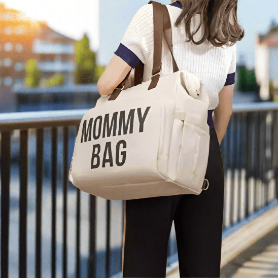 En plein air, près d'une balustrade, une femme porte le Sac a Langer | Mommy Bag - un sac à langer spacieux de couleur beige avec des poignées foncées et une inscription noire en gras "MOMMY BAG" - un accessoire essentiel pour les parents et leur bébé.