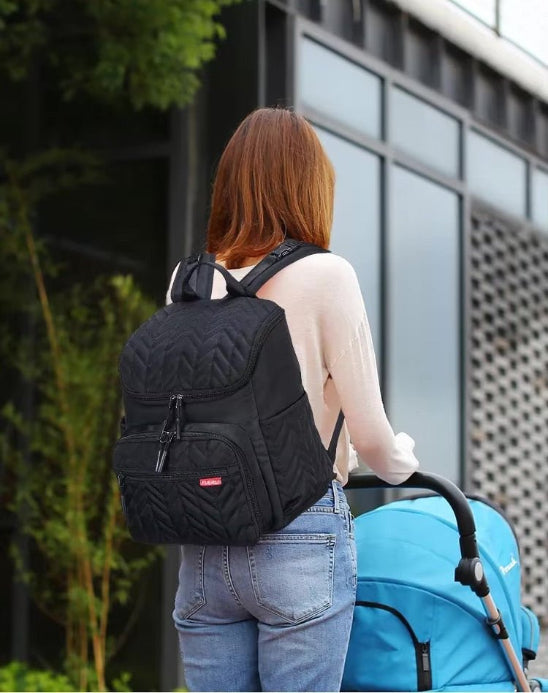 Une personne aux longs cheveux bruns, vêtue d'un pull léger et d'un jean, se tient en plein air, portant le Sac Bebe à Langer - NappyBag a dos et poussant une poussette bleue, avec de la verdure et un bâtiment moderne en arrière-plan.