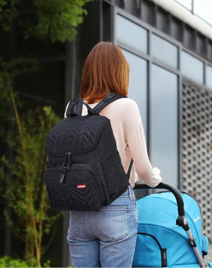 Une personne aux longs cheveux bruns, vêtue d'un pull léger et d'un jean, se tient en plein air, portant le Sac Bebe à Langer - NappyBag a dos et poussant une poussette bleue, avec de la verdure et un bâtiment moderne en arrière-plan.