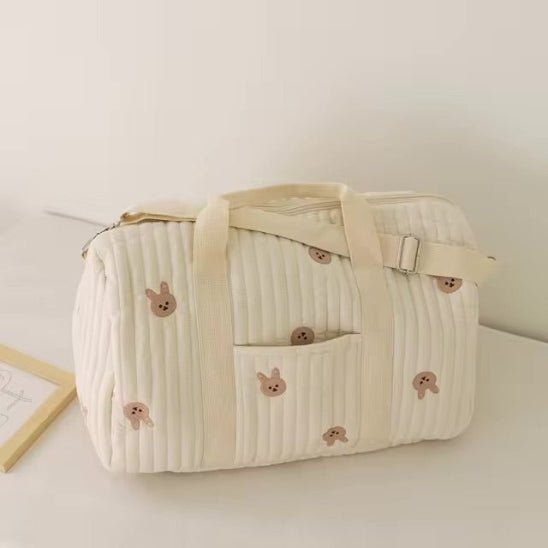 Un Sac bébé à Langer | Longchamp Bandoulière matelassé crème avec des poignées et une sangle beiges, représentant de petits visages d'animaux de bande dessinée marron, repose sur une surface blanche à côté d'un tableau encadré.