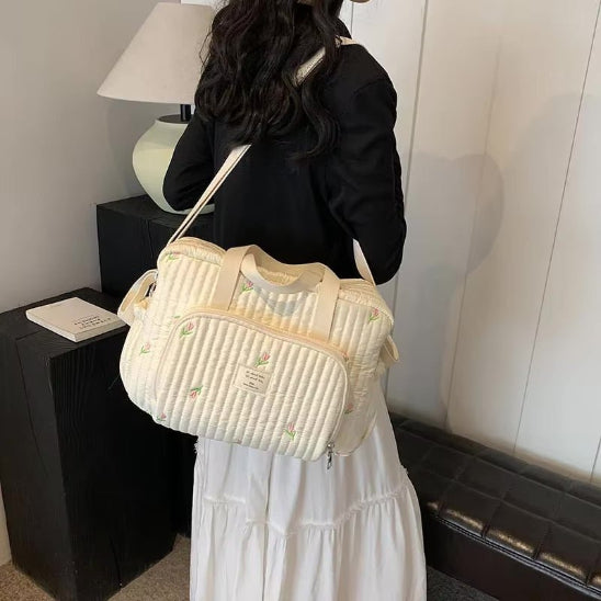 Une femme aux longs cheveux noirs, vêtue d'un haut noir et d'une jupe blanche, se tient debout à l'intérieur, tenant sur son épaule un Sac bebe à Langer | Longchamp Bandoulière avec des détails brodés. Une lampe et un livre sont posés sur une table noire à côté d'elle.