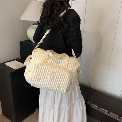 Une femme aux longs cheveux noirs, vêtue d'un haut noir et d'une jupe blanche, se tient debout à l'intérieur, tenant sur son épaule un Sac bebe à Langer | Longchamp Bandoulière avec des détails brodés. Une lampe et un livre sont posés sur une table noire à côté d'elle.