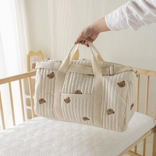 Une main tient le Sac bébé à Langer | Maternité, de couleur crème avec de jolis visages d'ours, au-dessus d'un berceau en bois et d'un matelas blanc - un choix idéal pour les mamans en mouvement dans une pièce doucement éclairée.