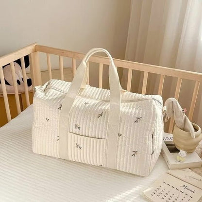 Un Sac bebe à Langer | Longchamp Bandoulière blanc matelassé avec des motifs de petites feuilles repose sur le matelas d'un berceau, à côté d'un panier de peluches et d'un calendrier, le tout dans une pièce à l'éclairage tamisé.