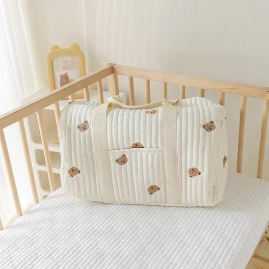 Un Sac bebe à Langer | Longchamp Bandouilère de couleur crème avec de petits visages d'ours bruns repose sur un matelas blanc dans un berceau en bois, tandis qu'une illustration d'ours encadrée se trouve sur une étagère à l'arrière-plan.