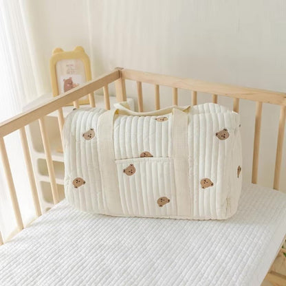Un Sac bebe à Langer | Longchamp Bandouilère de couleur crème avec de petits visages d'ours bruns repose sur un matelas blanc dans un berceau en bois, tandis qu'une illustration d'ours encadrée se trouve sur une étagère à l'arrière-plan.