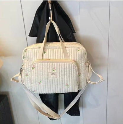 Un bandouilère matelassé Sac bebe à Langer | Longchamp crème avec de petits imprimés floraux et des bretelles beiges est suspendu à un crochet au-dessus d'une veste noire contre un mur clair.