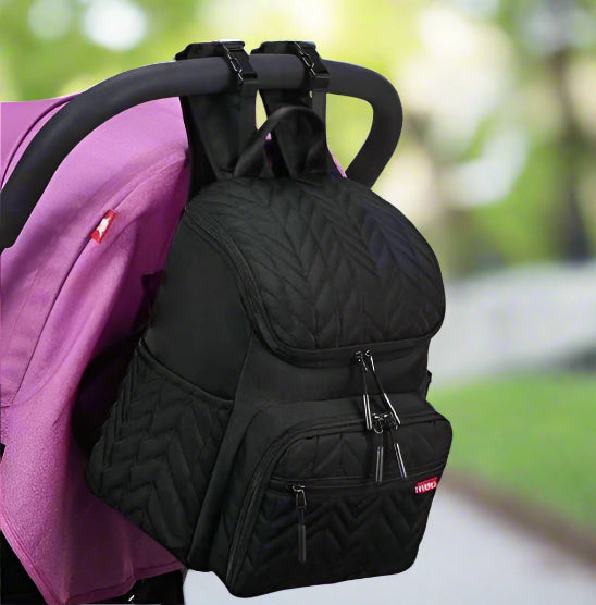 Le Sac Bebe à Langer - Nappy, avec son motif matelassé noir et ses bretelles réglables, pend élégamment du guidon dune poussette violette. Il offre plusieurs compartiments zippés pour une combinaison parfaite délégance et de fonctionnalité lors de vos sorties.