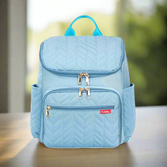 Le Sac Bebe à Langer - NappyBag a dos, un sac à dos matelassé bleu clair avec deux compartiments zippés, deux poches latérales et une poignée bleue, repose sur du bois. La poche avant est ornée d'une étiquette FUERLI rouge avec texte blanc.